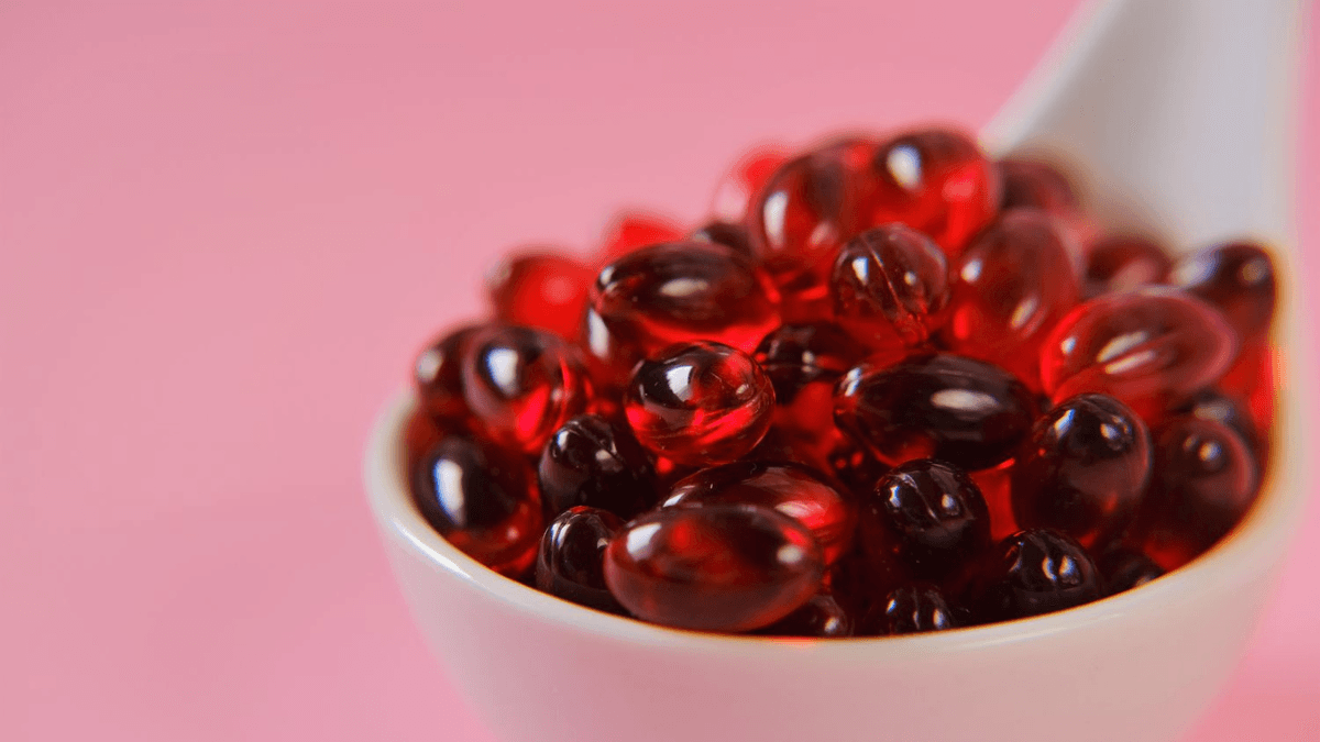 Close-up of red krill oil softgel capsules in a white spoon against a pink background.