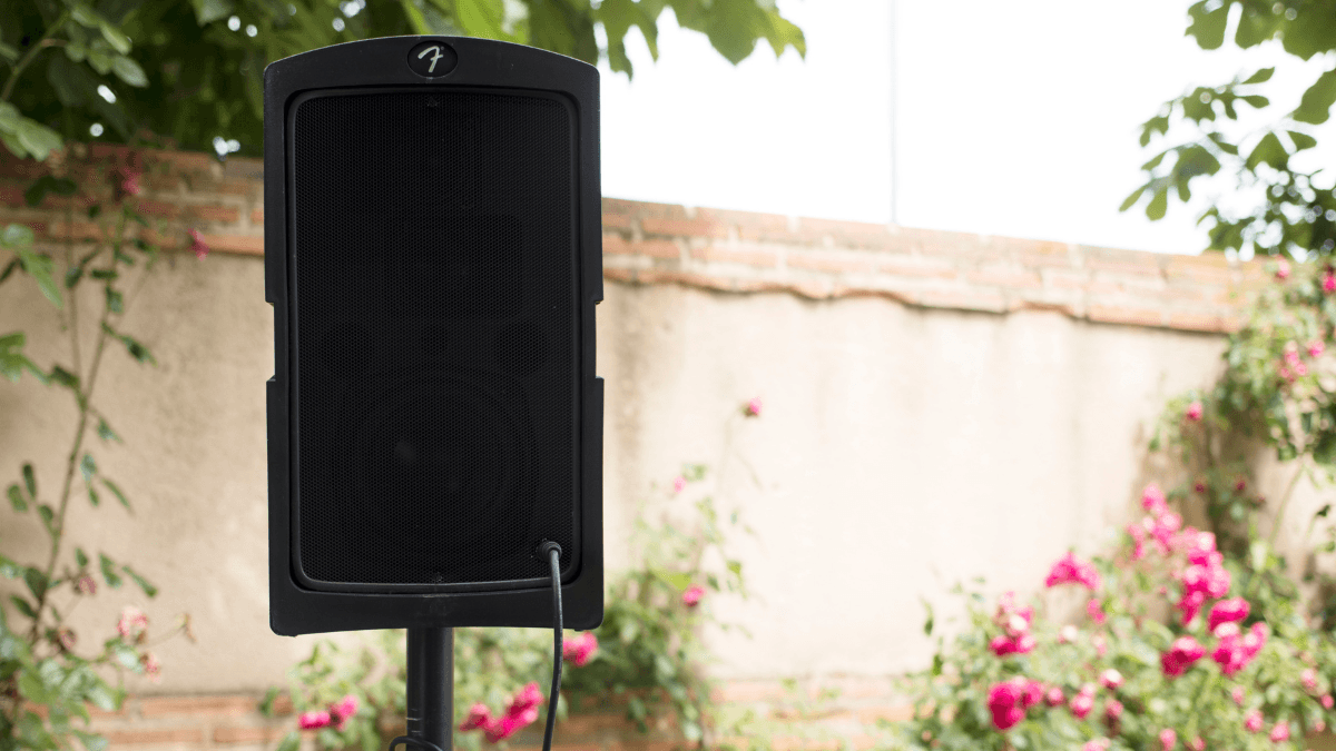 A black outdoor speaker on a stand is set up in a garden with flowers and a brick wall in the background.