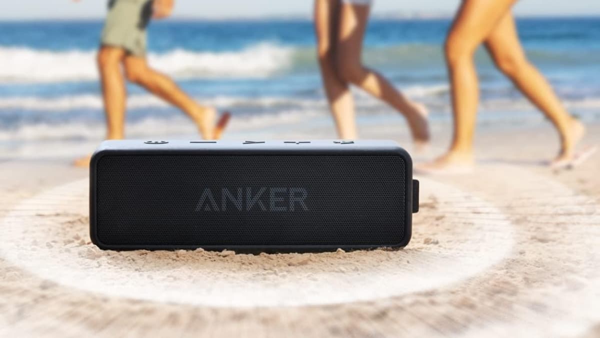 Anker Soundcore 2 portable Bluetooth speaker sitting on sand with people walking by the beach.
