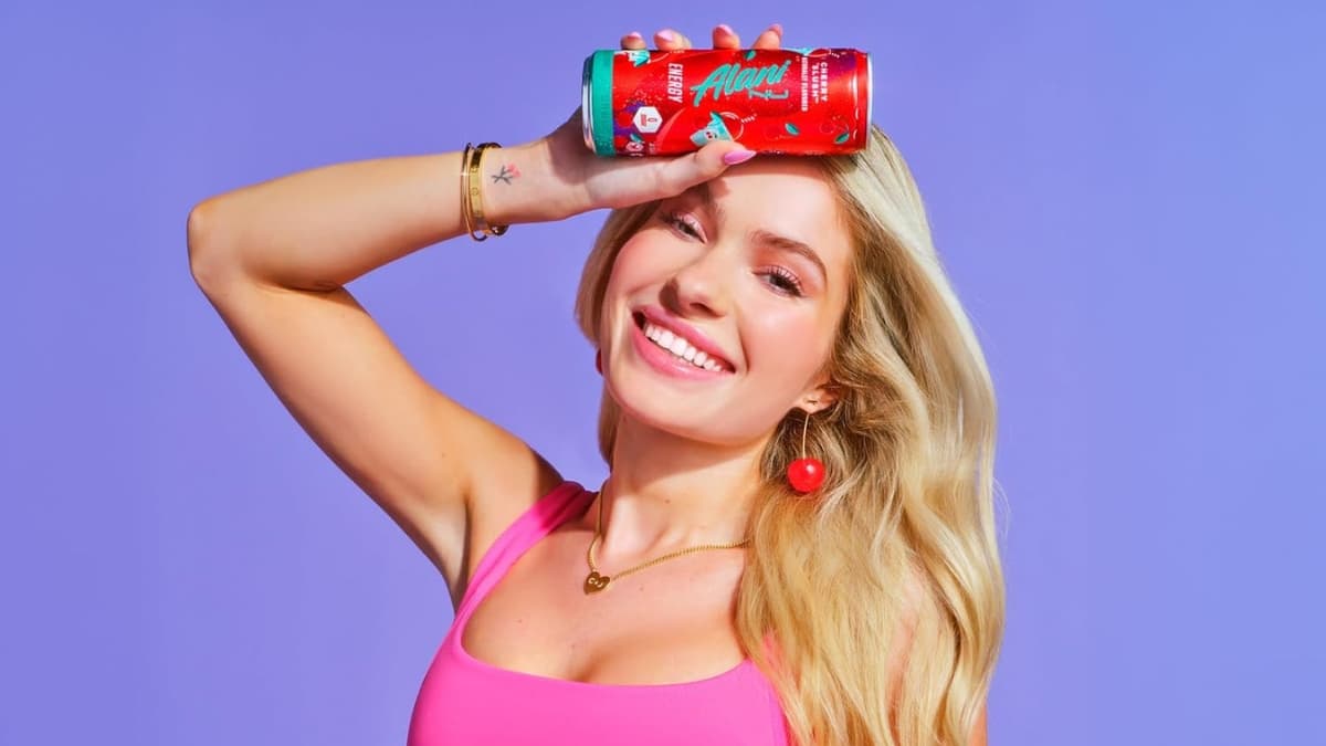Smiling woman in a pink top holds an Alani Nu Cherry Slush Energy Drink can against her forehead.