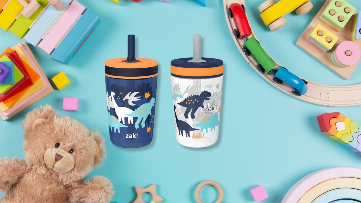 Two Zak Designs Kelso Toddler Cups with dinosaur designs sitting on a playroom table with toys.